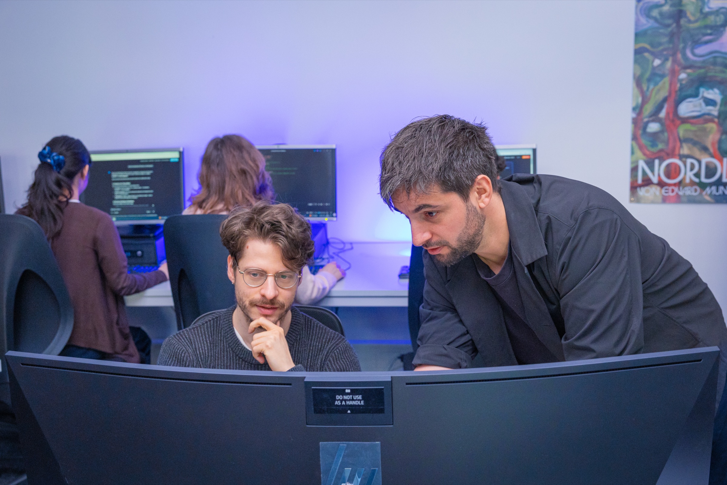 Duo dev: mentor à droite penche sur l’écran, halo violet sur le mur